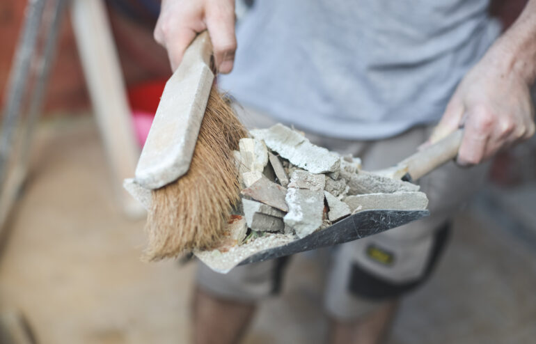 A young Caucasian man holds a scoop with construction waste and a broom to throw it into a bag